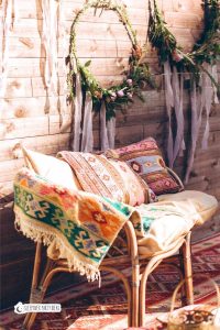 Boho graduation party ideas with bohemian-style seating area with a rattan bench adorned with colorful patterned pillows and a woven blanket, set against a wooden backdrop decorated with floral wreaths and ribbons